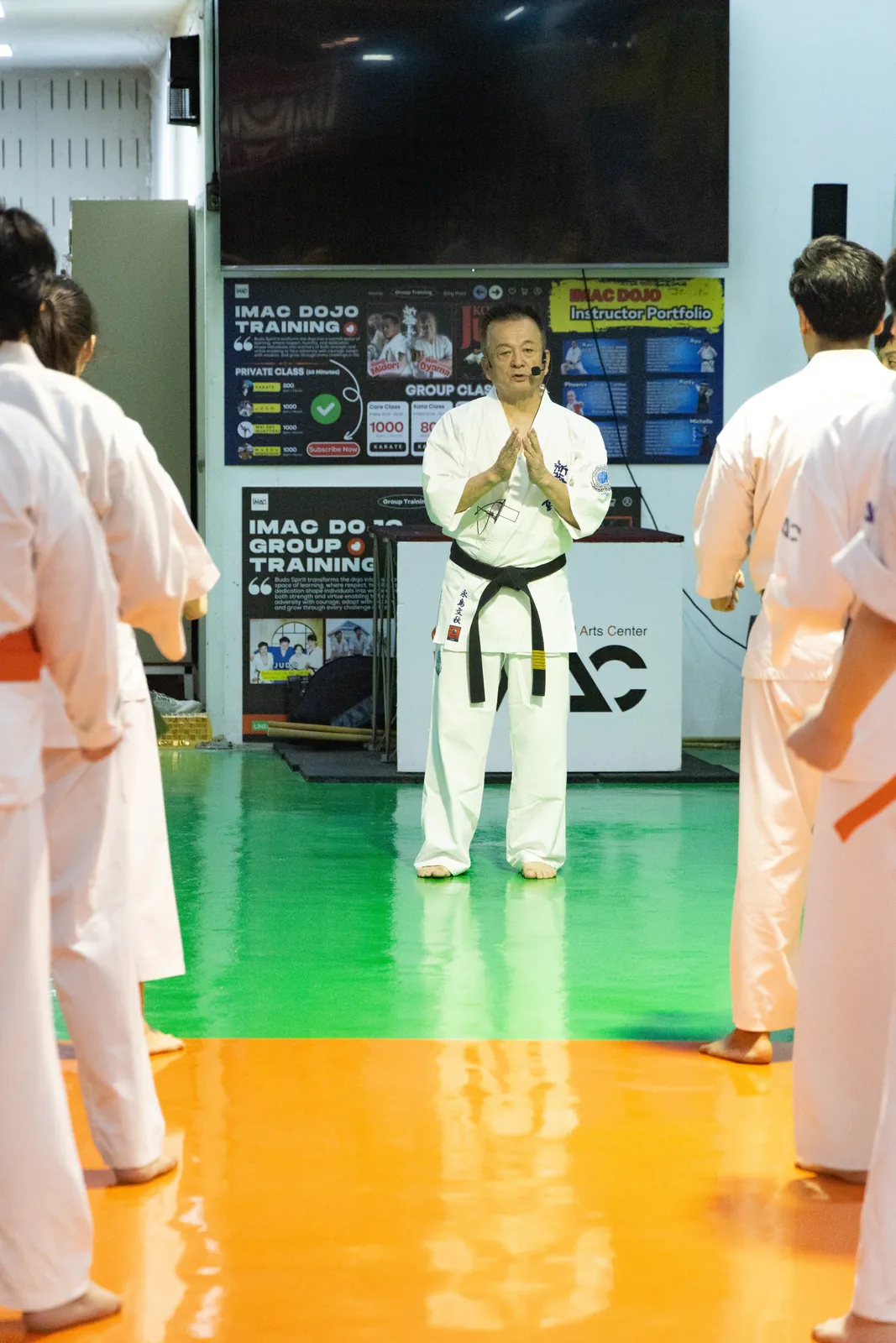 Shihan Nagashima ยืนพนมมือทักทายนักเรียนที่ยืนเป็นแถวสองข้างในโดโจ