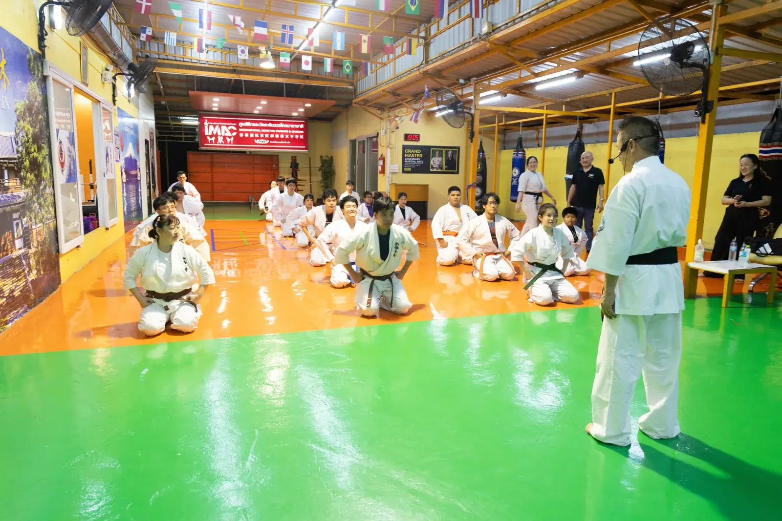 นักเรียนคาราเต้นั่งคุกเข่าเป็นแถวบนพื้นสีส้ม หันหน้าเข้าหา Shihan ที่ยืนอธิบายด้านขวา