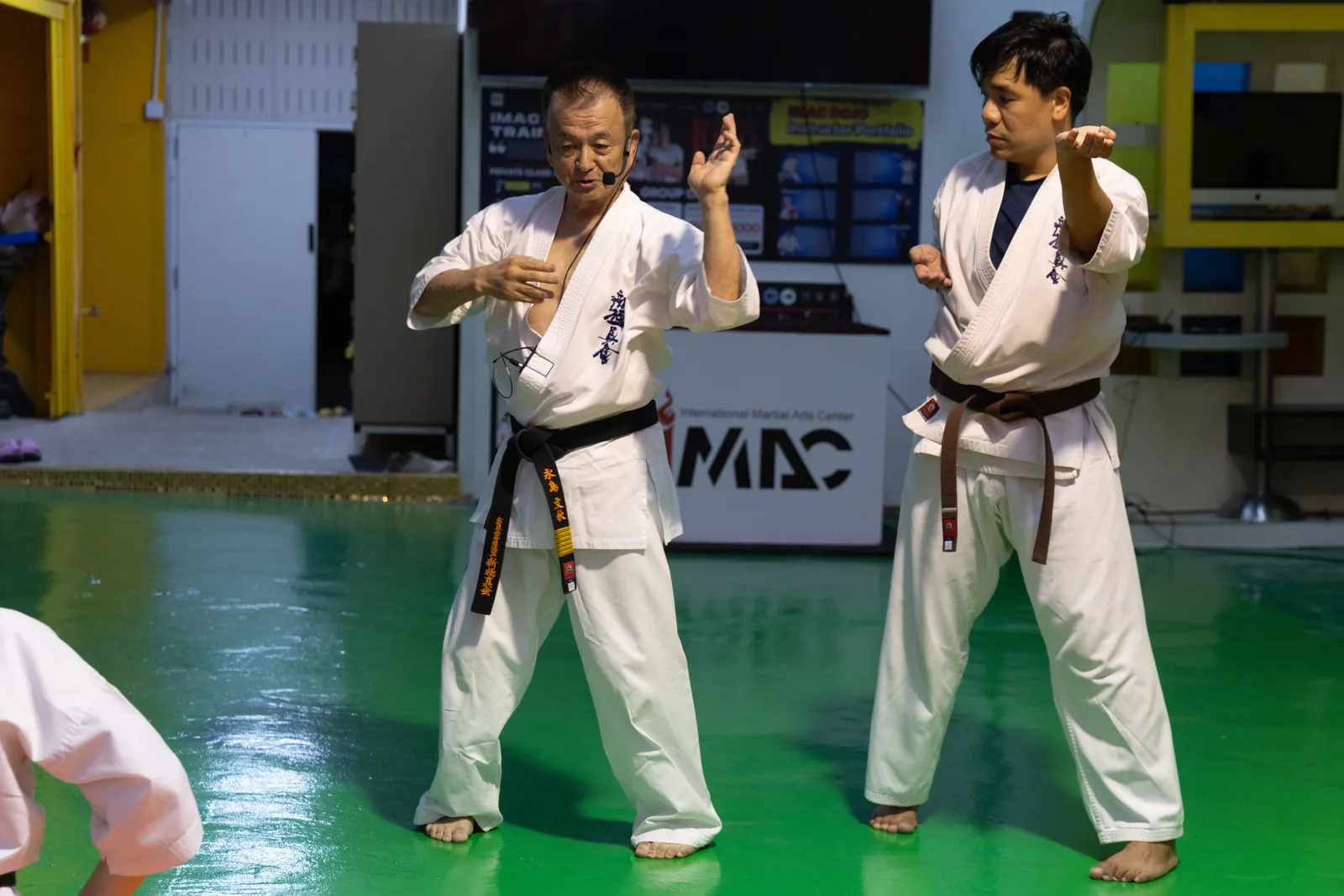 Shihan Nagashima ยกมือทั้งสองข้างอธิบายจังหวะ ขณะครูสายน้ำตาลยืนเตรียมท่าอยู่ด้านขวา