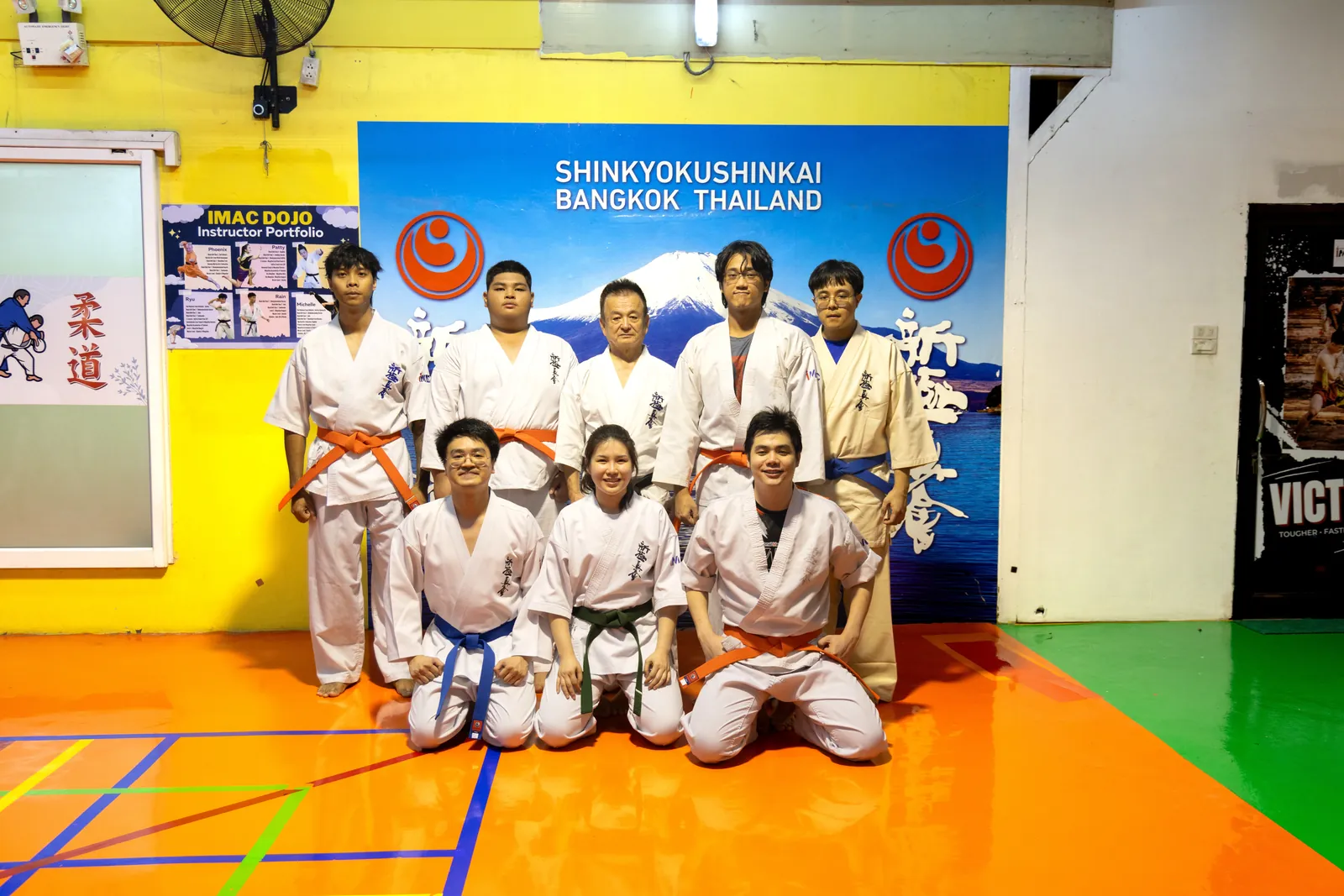 ผู้รับสายแปดคนถ่ายภาพร่วมกับ Shihan Nagashima หน้าฉาก Shinkyokushinkai Bangkok Thailand มีสายส้ม น้ำเงิน และเขียว