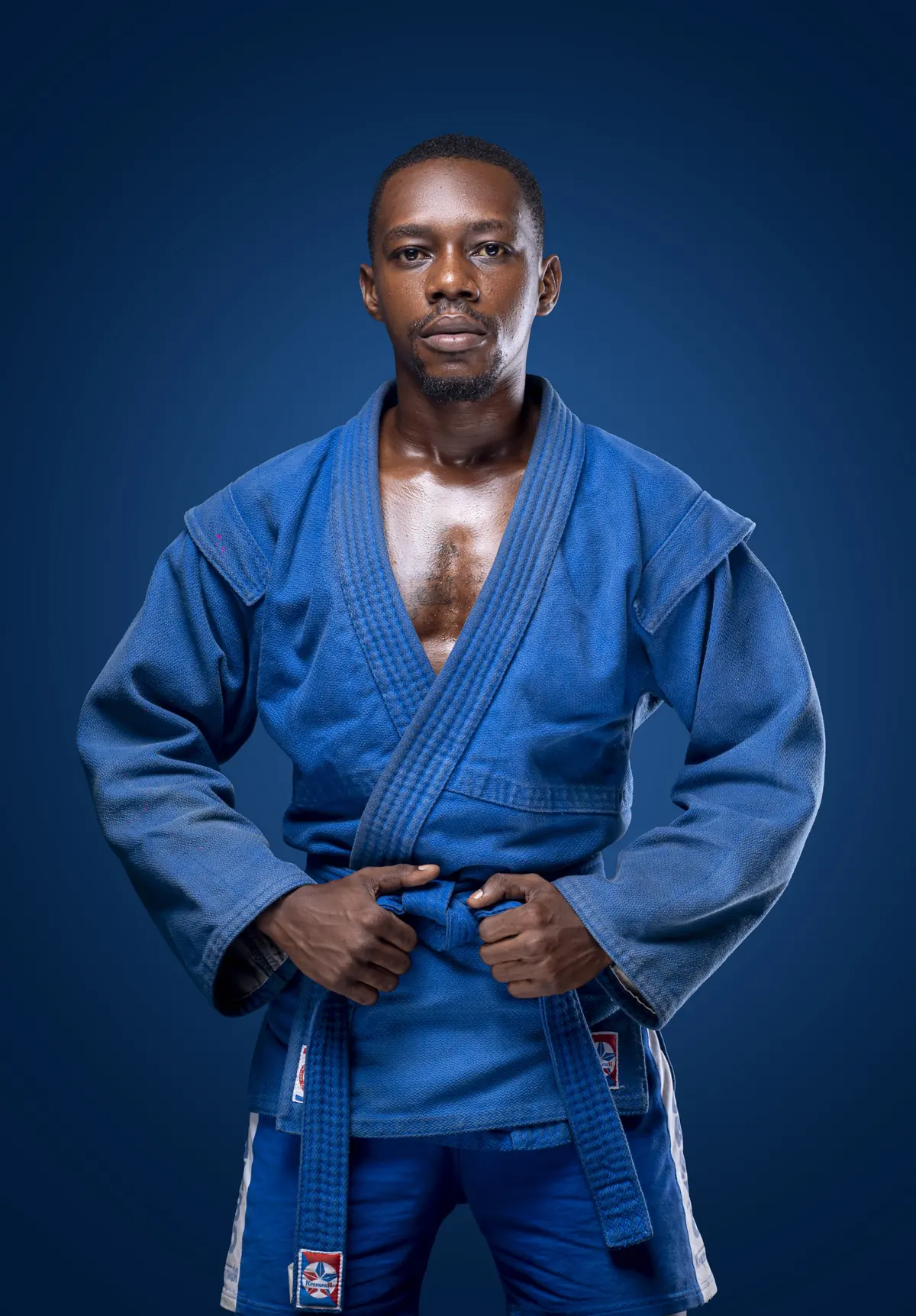 A man in a blue sambo jacket with a belt standing with hands clasped in front, against a dark blue background