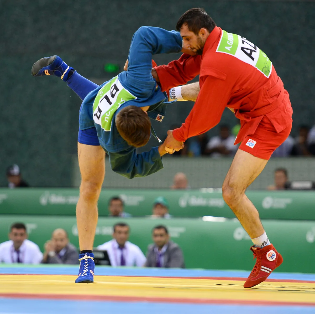 A sambo competitor in red lifting an opponent in blue off the mat in a throw