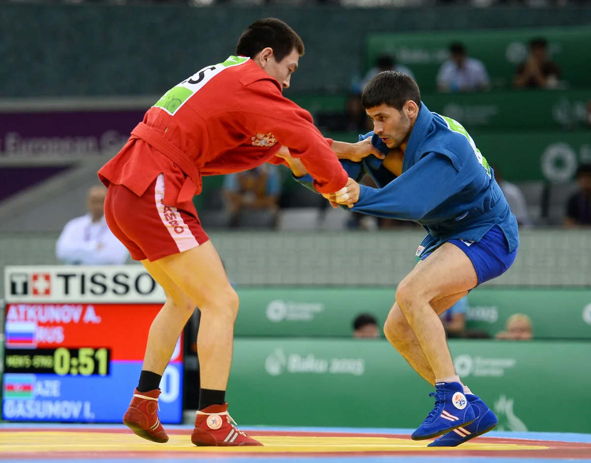 Sambo fighters in red and blue gripping each other at close range on the mat, with a scoreboard on the left