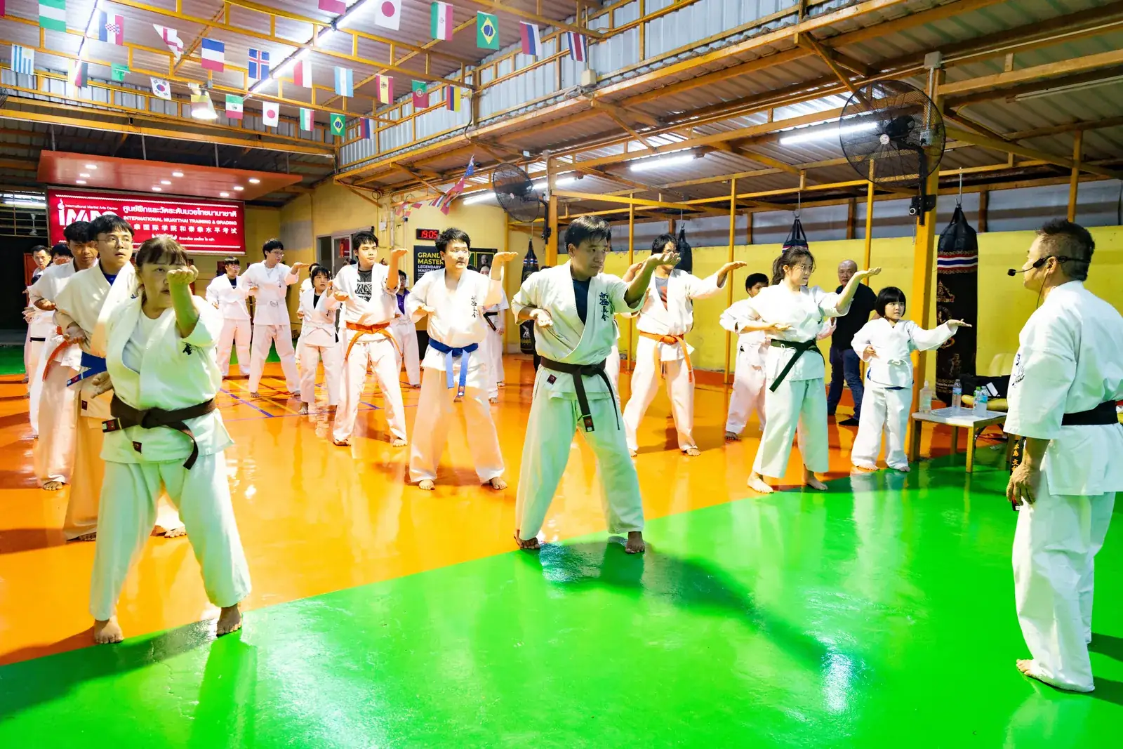 Shihan Fumiaki Frank Nagashima ยืนสังเกตนักเรียนทั้งคลาสที่กำลังฝึก Kihon บนพื้นสีเขียวและส้มของ IMAC Dojo